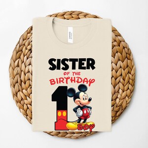 May include: A beige t-shirt with the text "SISTER OF THE BIRTHDAY" in black and red. A large number "1" is next to a cartoon image of Mickey Mouse. The shirt is folded on a woven, circular mat.