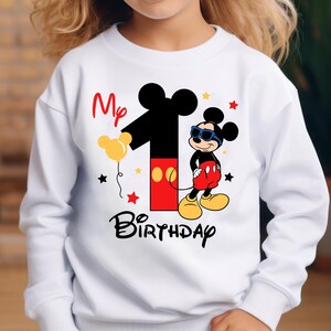 May include: White sweatshirt featuring a graphic of Mickey Mouse wearing sunglasses, with the number 1 and the words "My Birthday". The design includes yellow, red, and black elements, with star accents.