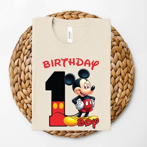 May include: A cream-colored t-shirt featuring a graphic of Mickey Mouse with the text "BIRTHDAY BOY" and the number "1" in red and black. The shirt is displayed on a woven, circular placemat.