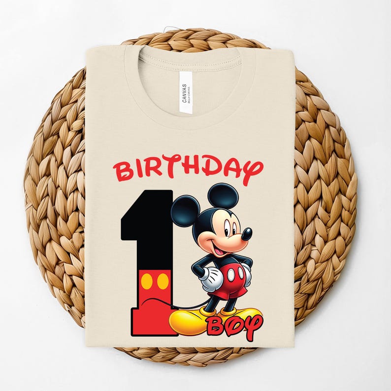 May include: A cream-colored t-shirt featuring a graphic of Mickey Mouse with the text "BIRTHDAY BOY" and the number "1" in red and black. The shirt is displayed on a woven, circular placemat.