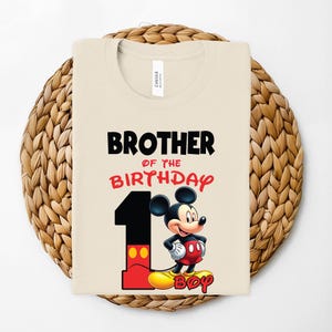 May include: A beige t-shirt with the text "BROTHER OF THE BIRTHDAY" in black and red. A large number "1" is on the left, with Mickey Mouse standing next to it. The word "BOY" is at the bottom in yellow.