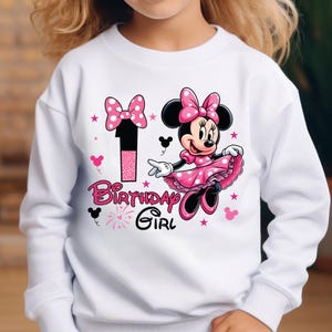 May include: White sweatshirt featuring Minnie Mouse in a pink polka-dot dress and bow. The number 1 and the words "Birthday Girl" are in pink. Black Mickey Mouse silhouettes and pink stars complete the design.