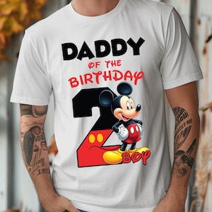 May include: White t-shirt with the text "DADDY OF THE BIRTHDAY BOY" in black and red. A graphic of Mickey Mouse is featured, along with the number 2 in black and red. The design is likely for a birthday celebration.