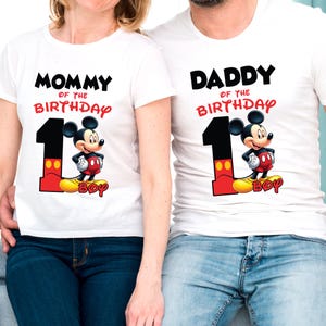 May include: White t-shirts with matching designs. One shirt reads "MOMMY of the BIRTHDAY" and the other reads "DADDY of the BIRTHDAY". Both shirts feature a large number "1" with Mickey Mouse and the word "BOY".