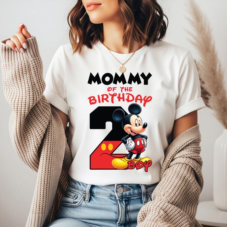 May include: White t-shirt with the text "MOMMY OF THE BIRTHDAY BOY" in black and red. A large number "2" is featured with Mickey Mouse. The shirt is a birthday theme.