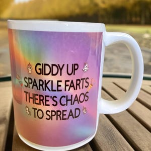 May include: White ceramic coffee mug with a rainbow ombre design and the text "Giddy Up Sparkle Farts There's Chaos To Spread". The mug has a white handle and is decorated with small unicorn and rainbow illustrations.