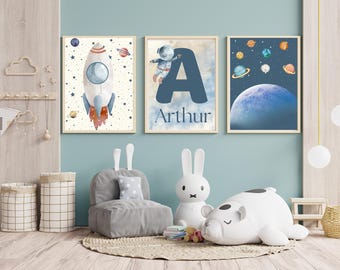 May include: Three framed prints with a space theme. The first print features a white rocket ship with orange flames against a white background with blue stars. The second print features a blue letter A with a white astronaut in a space suit. The text "Arthur" is below the letter. The third print features a blue planet with a white ring against a dark blue background with white stars and smaller planets.