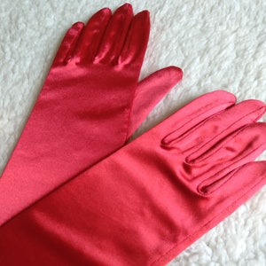 May include: A pair of long, red satin gloves. The gloves are folded in half, with the fingertips of one glove resting on the back of the other glove.