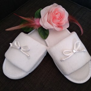 May include: A pair of white fuzzy slippers with satin bows and rhinestones. The slippers are on a dark gray surface with a pink rose and feathers.