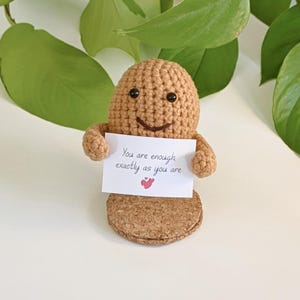 Puede incluir: Una figura de cacahuete de ganchillo que sostiene un cartel que dice "You are enough exactly as you are" con un pequeño corazón rojo dibujado debajo del texto. La figura está de pie sobre una base de corcho.