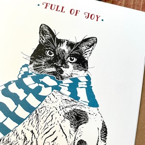 May include: A black and white cat wearing a blue and white striped scarf. The cat is looking at the camera. The text "FULL OF JOY." is written in red above the cat.