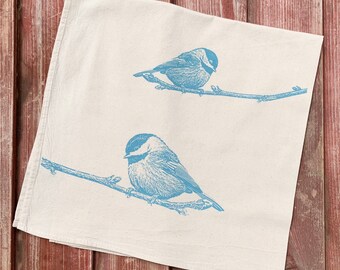Puede incluir: Una toalla de cocina blanca con una silueta azul de dos pájaros posados en ramas. Los pájaros tienen un cuerpo redondo y un pico pequeño.