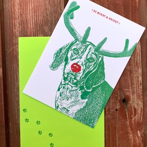 May include: A green and white Christmas card featuring a dog with antlers and a red nose. The card reads "(Be Merry & Bright)" and has green paw prints on the bottom.