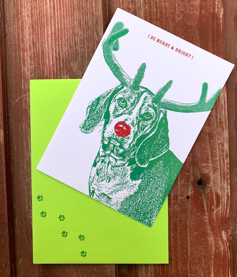 May include: A green and white Christmas card featuring a dog with antlers and a red nose. The card reads "(Be Merry & Bright)" and has green paw prints on the bottom.
