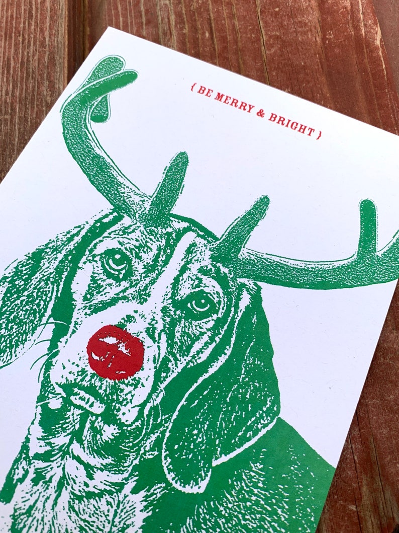 May include: A green and white illustration of a dog wearing a red nose and antlers. The text (BE MERRY & BRIGHT) is printed in red.