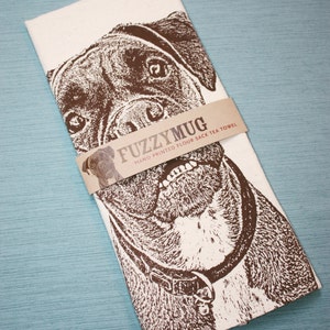 May include: A white tea towel with a brown and white illustration of a dog's face. The text "Fuzzy Mug" is printed on a brown label across the image. The label also says "Hand Printed Flour Sack Tea Towel".