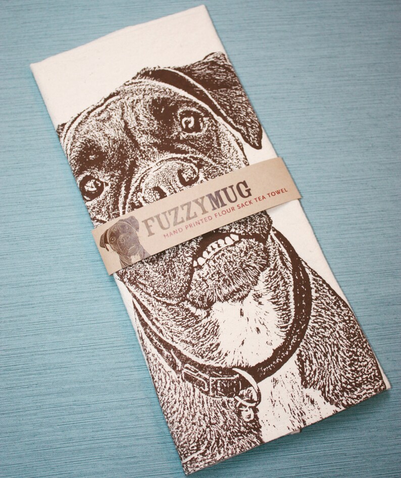 May include: A white tea towel with a brown and white illustration of a dog's face. The text "Fuzzy Mug" is printed on a brown label across the image. The label also says "Hand Printed Flour Sack Tea Towel".