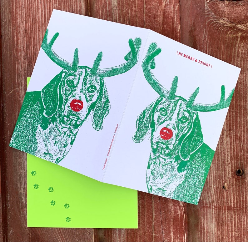 May include: A Christmas card featuring a green illustration of a dog wearing reindeer antlers with a red nose. The text on the card reads "(BE MERRY & BRIGHT)".