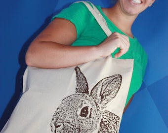 May include: A white canvas tote bag with a brown rabbit illustration. The bag has a green strap and is being held by a person.