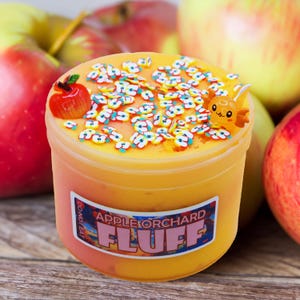 May include: A container of yellow-orange slime with apple-shaped confetti and a small orange character. The container has a label that reads "Apple Orchard Fluff". A red apple is on top of the container. Apples are in the background.