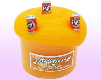May include: Orange-colored slime in a clear container with a label that reads "Mell-Orange Pop DO NOT EAT." Three miniature Fanta soda cans are on top of the slime. The background is a light purple.