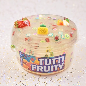 May include: A clear container of slime with fruit-themed decorations. The slime is filled with colorful fruit slices and topped with strawberry, pineapple, and pear charms. The container has the words "TUTTI FRUITY" on the front.