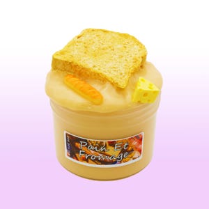May include: A jar of yellow slime with a slice of toast, a baguette, and a cheese wedge on top. The jar has a label with the words "Pain Et Fromage". The background is a gradient of white to light purple.