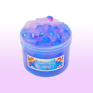 May include: A clear plastic container filled with blue slime, topped with clear and blue beads. The container has a label that reads "MERMAID BUBBLES" in colorful text. The slime has a translucent appearance.