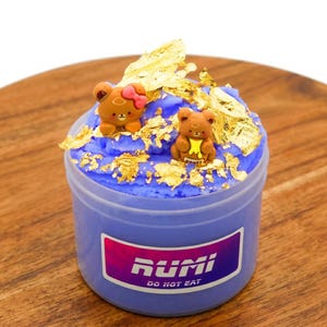 May include: A small, clear plastic container filled with a vibrant blue substance, topped with gold leaf and two small bear figurines. The container has a label with the text "RUMI DO NOT EAT". The container sits on a wooden surface.