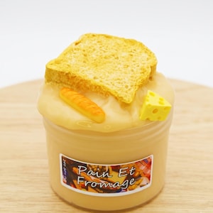 May include: A clear plastic container filled with light tan slime, topped with a miniature yellow slice of bread, a small baguette, and a yellow cheese wedge. The label on the front features a colorful food graphic and the text Pain Et Fromage.