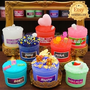 May include: A collection of colorful, decorative slime containers with various toppings and labels. The slimes are in shades of blue, red, pink, and green, with decorative elements like hearts, stars, and small figurines. The containers are displayed on ornate golden stands.