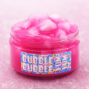May include: A jar of pink slime with the words "Bubble Bubble Yum" on a label. The slime has a bubbly texture and is a vibrant pink color. The jar is clear with a pink base.