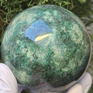 May include: A spherical mineral specimen featuring a textured surface with variegated shades of deep forest green, emerald, and dark teal. The stone displays a polished, reflective patch near the center that catches light, revealing a smooth, glossy finish.
