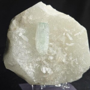May include: A light green aquamarine crystal specimen. The main crystal is a pale blue-green color, with a rectangular shape. It is set on a matrix of white and light green minerals. The background is black.