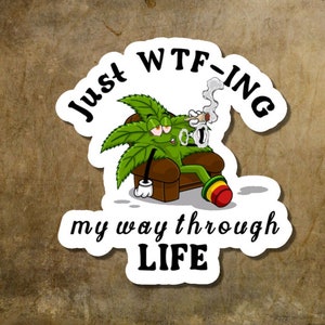 May include: A sticker featuring a cartoon cannabis leaf character relaxing in a brown armchair, smoking a cigarette. The sticker has the text "Just WTF-ing my way through LIFE" in a playful font. The background is a textured, brown surface.