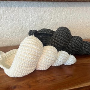 May include: Crocheted seashell sculptures in white and dark gray. The white shell is in the foreground, with a spiral design. The dark gray shell is behind it. Both are on a wooden surface.