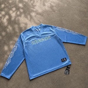 May include: Light blue long-sleeve jersey with the word "CHROME" in a stylized font across the chest. The sleeves feature white decorative patterns. A tag with a logo hangs from the bottom hem.