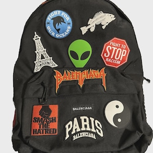 Könnte beinhalten: Schwarzer Rucksack mit verschiedenen Aufnähern. Aufnäher zeigen einen grünen Alienkopf, ein Fischskelett, ein rotes Stoppschild mit der Aufschrift "Fight to Stop Racism" und einen Aufnäher mit "Smash the Hatred". Der Rucksack trägt auch das Wort "Balenciaga".