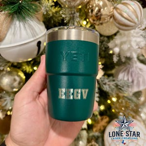 May include: A teal Yeti tumbler with the brand name embossed on the front. The bottom of the tumbler has the letters EEGV engraved. The tumbler has a silver rim and is held in front of a Christmas tree.