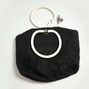 May include: A black beaded handbag with a white handle. The bag has a rounded shape and a white circular handle attached with a silver-colored metal clasp. The bag's surface is covered in small, raised beads.