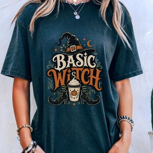 May include: A dark charcoal gray short-sleeve t-shirt featuring a graphic print with the text "BASIC WITCH" in cream and orange. The design includes a black witch hat, a coffee cup with a pumpkin icon, and two black boots with striped socks.