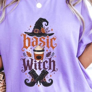 May include: A light purple t-shirt featuring a graphic print with a black witch hat, a coffee cup with a pumpkin icon, and black boots. The text "basic Witch" is printed in orange and dark purple lettering, surrounded by small star and leaf accents.