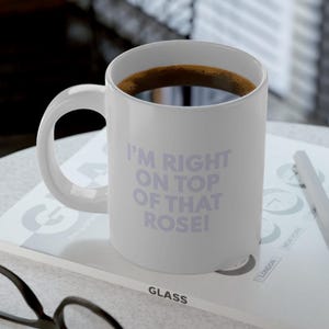 Puede incluir: Taza de café de cerámica blanca con asa negra. La taza tiene el texto "I'm right on top of that rose!" impreso en color morado claro.