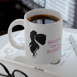 Può includere: Tazza da caffè in ceramica bianca con una silhouette nera di una donna con una coda di cavallo. Il testo "Baddie on a Budget" è stampato in rosa sulla tazza.