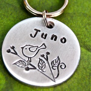 May include: Silver dog tag with a small bird and leaves engraved on it. The tag says "Juno" on it.