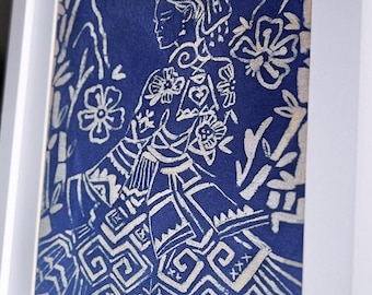 May include: Framed artwork featuring a blue fabric print with a white design of a figure in traditional attire. The artwork is framed in a white, square frame. The print includes floral and geometric patterns.