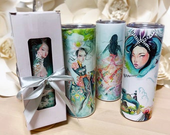 May include: Three stainless steel tumblers with colorful illustrations of women in traditional Asian clothing. The tumblers are on a wooden surface with white paper flowers in the background. One tumbler is in a white gift box with a silver ribbon.