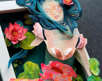 May include: A framed paper art piece depicting a serene woman with closed eyes, surrounded by water lilies. The artwork features a color palette of pink, blue, and green, with detailed paper cutouts of the woman, flowers, and lily pads.