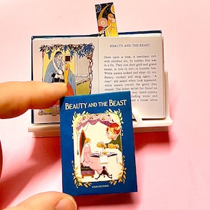 Peut inclure: Un petit livre bleu intitulé "Beauty and the Beast" avec une illustration colorée sur la couverture. Le livre est ouvert, révélant du texte et des illustrations. Un marque-page avec un design céleste est visible. Le thème est les contes de fées.