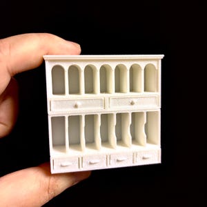Puede incluir: Un pequeño mueble blanco en miniatura con aberturas arqueadas, cajones y estantes. El mueble es una réplica detallada, ideal para casas de muñecas o exhibiciones en miniatura. El mueble es sostenido por una mano sobre un fondo negro.